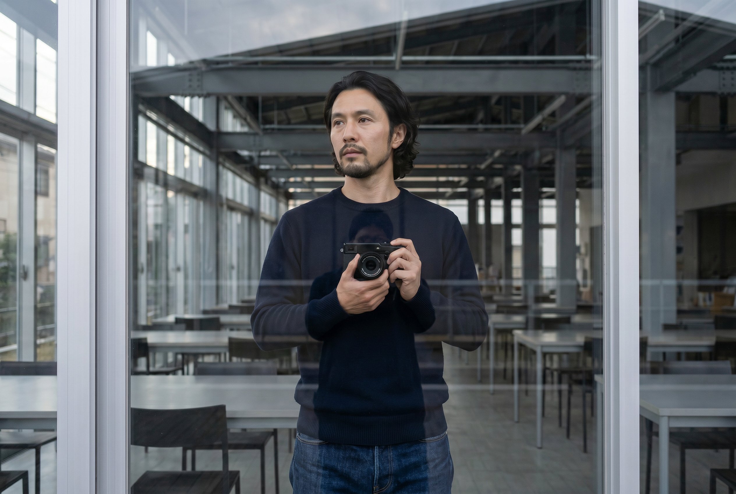 Partial reflection in the full-height glazed front facade, a figure standing on the side street outside with Fuji X-Pro3 raised, behind the reflection the interior reading tables and exposed girders visible through the glass