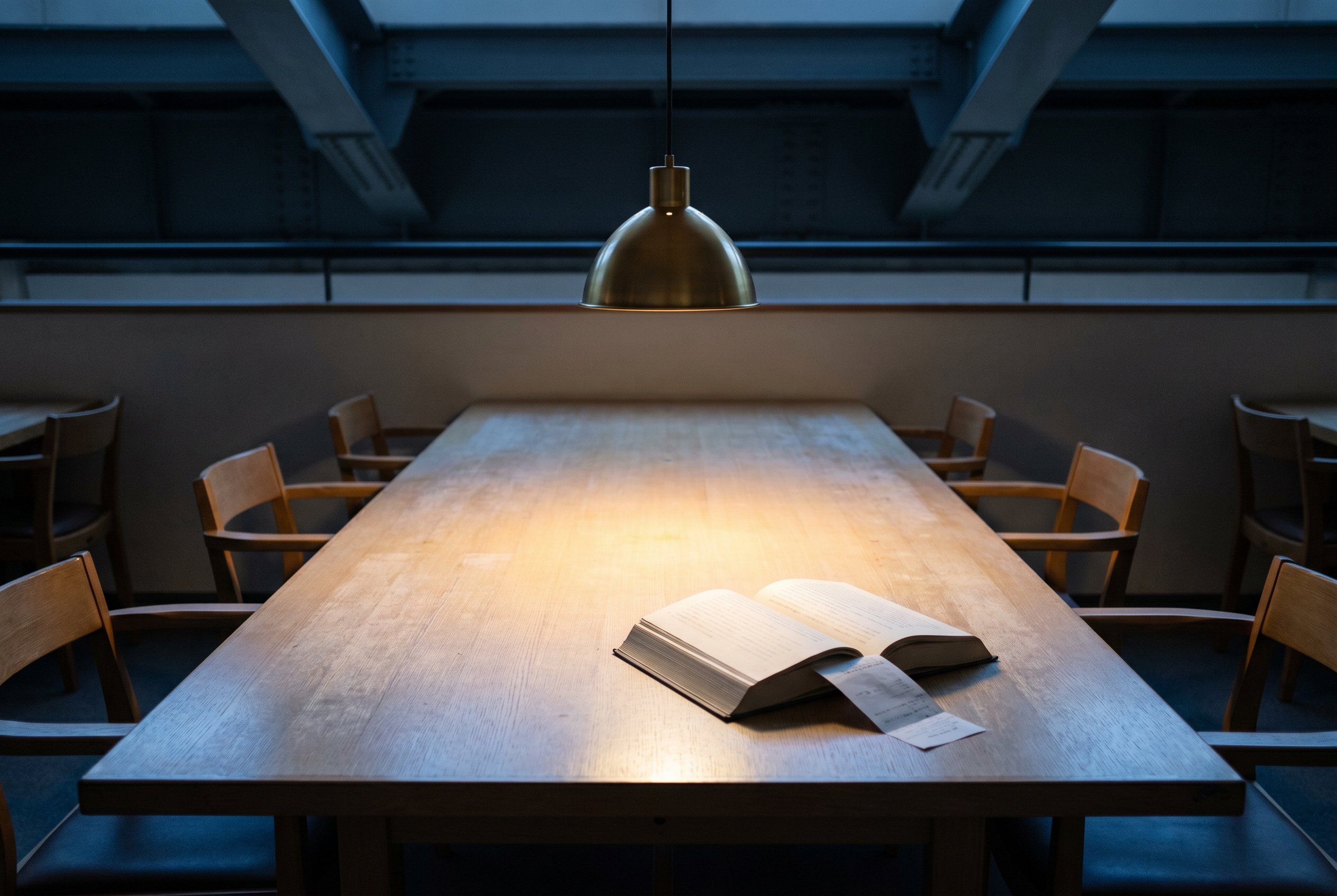 An empty pale-oak reading table with a single open hardback book lying face-down, a low brass pendant lamp throwing a focused warm pool of light onto the table surface