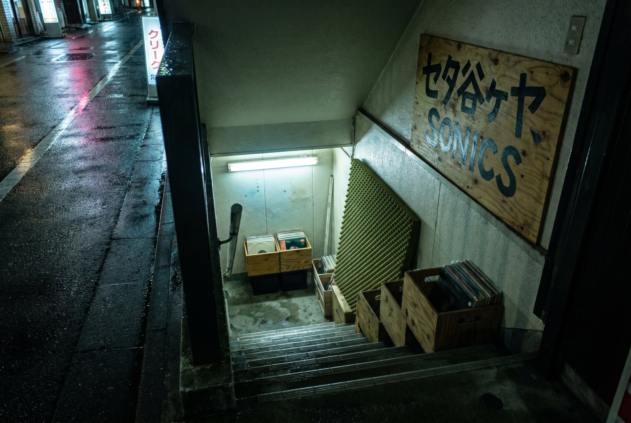 Looking down a narrow concrete staircase from sidewalk level into the shop, ten steps descending, fluorescent tube at the base